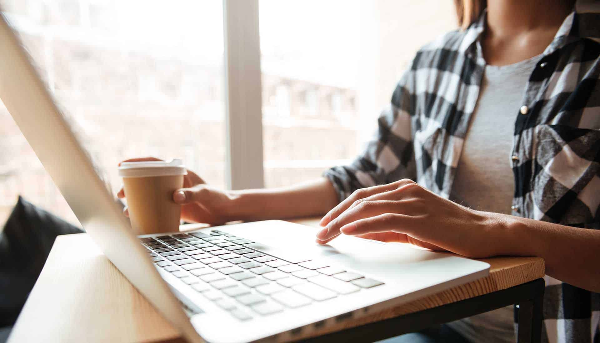 Cropped photo of attractive young caucasian lady sitting indoors using laptop computer and drinking coffee.