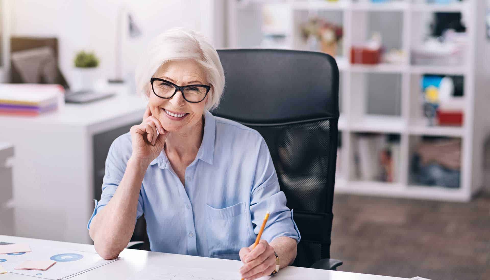 Happy charming aged businesswoman sitting at the table in front of the tablet in the office.